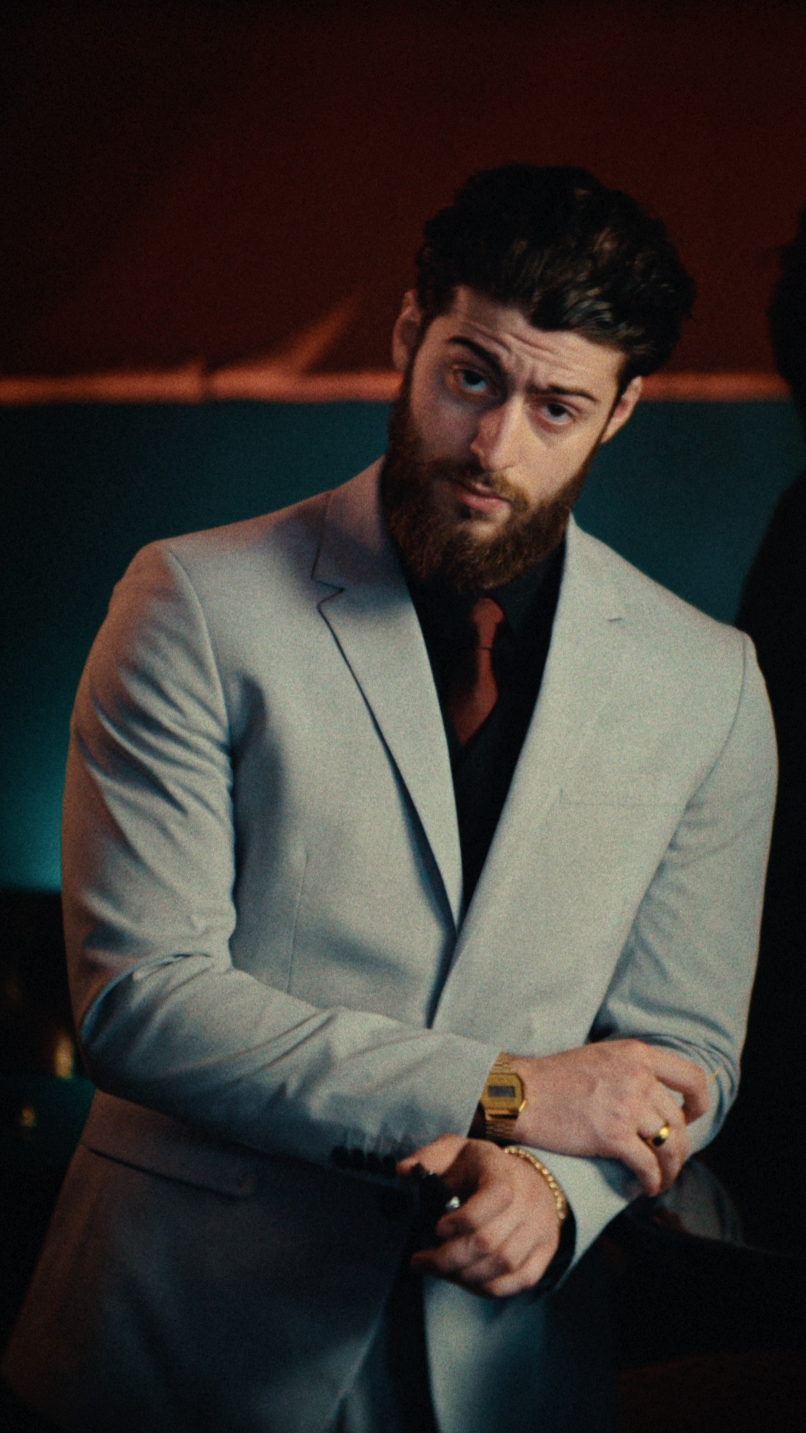 Un homme barbu en costume gris clair ajuste sa manche, une montre dorée au poignet, devant un mur partagé entre orange et turquoise.