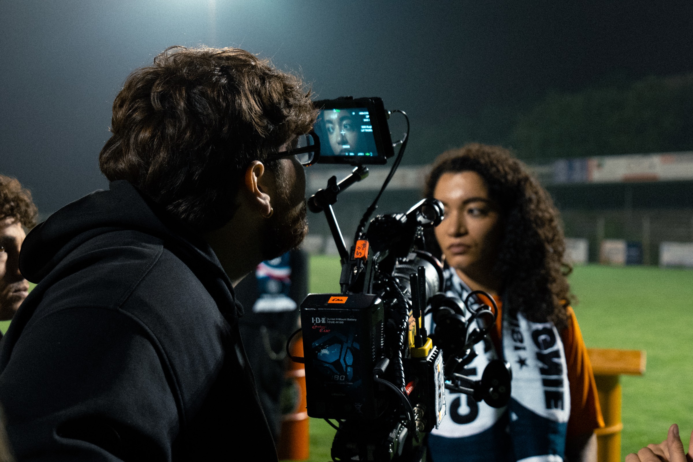 Youen Frémont à l'œilleton d'une caméra épaule, lors d'un tournage de nuit sur un terrain de football éclairé ; une jeune femme en maillot de supporter au second plan.