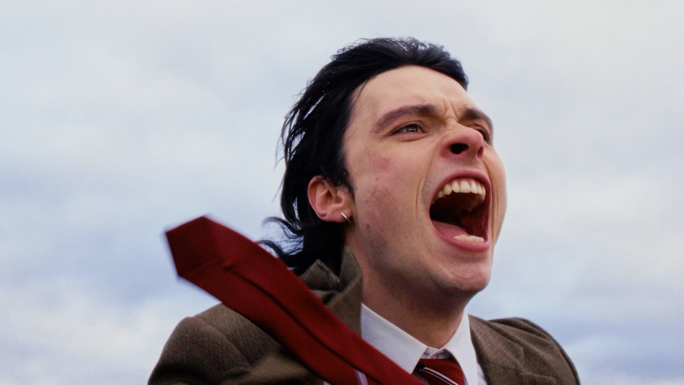 Un homme en costume brun et cravate rouge crie, bouche grande ouverte, la cravate soulevée par le vent devant un ciel nuageux.