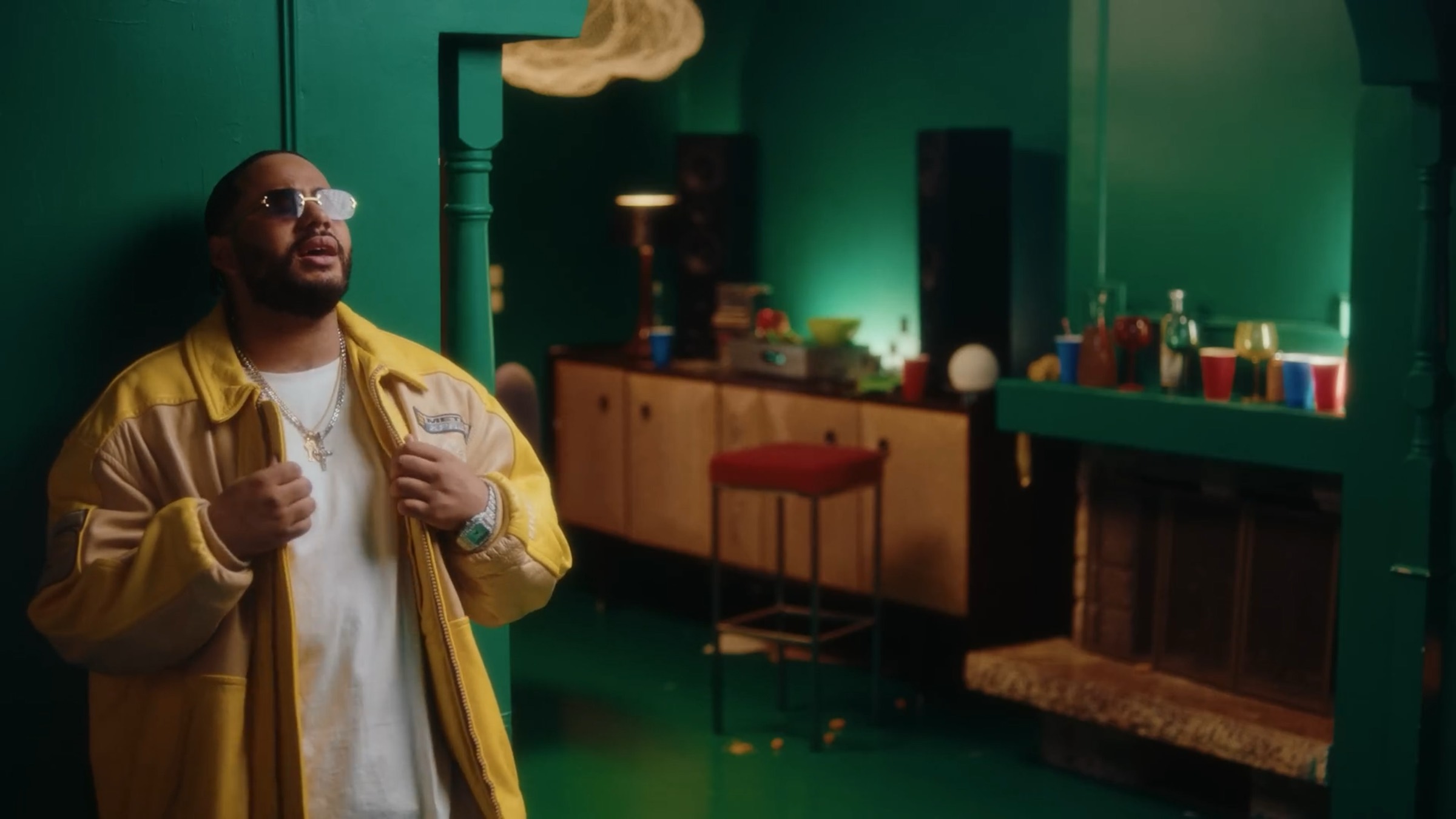 Un homme en veste jaune ouverte sur un t-shirt blanc chante dans un salon vert, un bar et des bouteilles derrière lui.