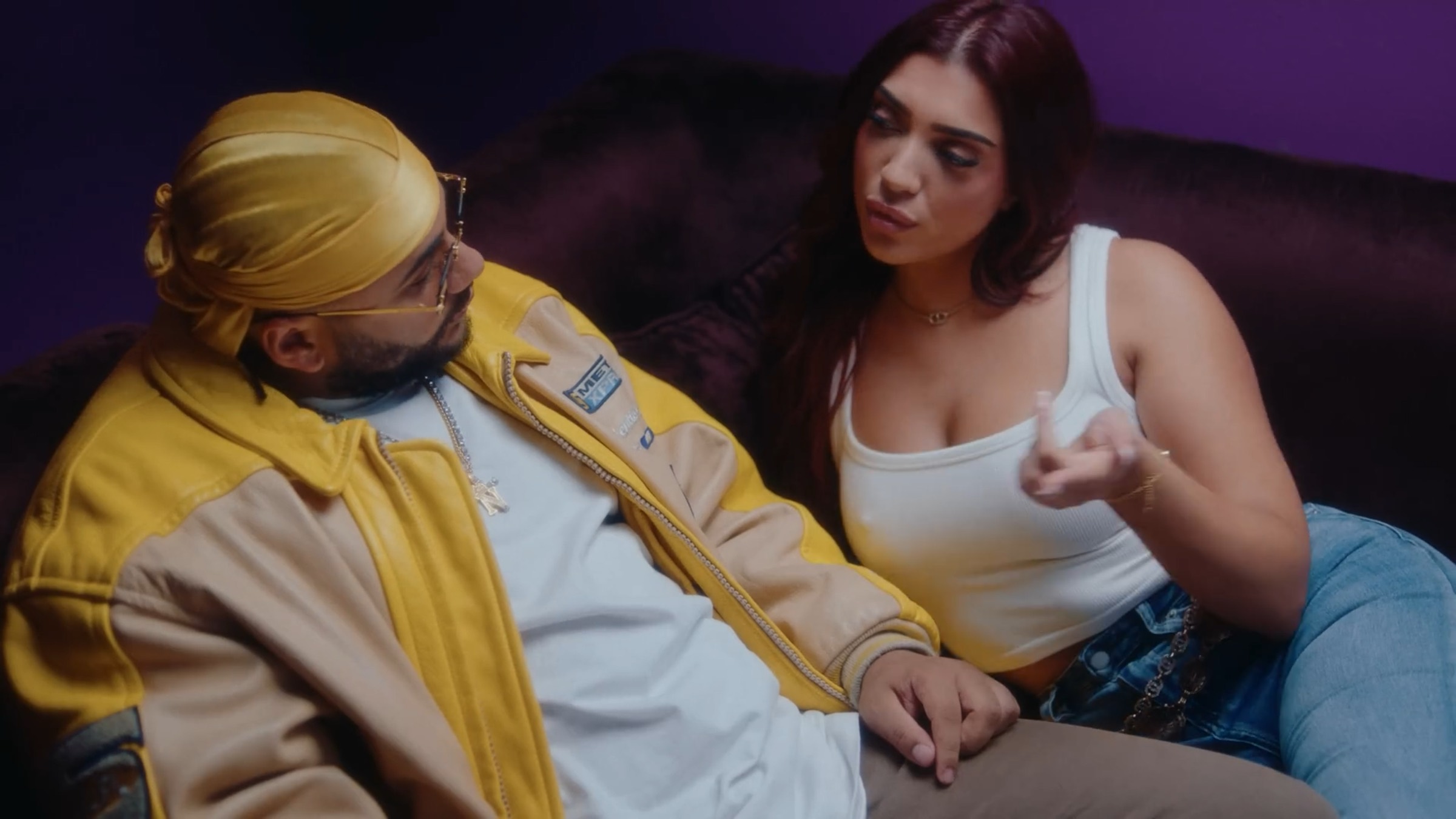 Un homme en durag jaune assis sur un canapé, une jeune femme en débardeur blanc lui parle de côté, mur violet derrière.