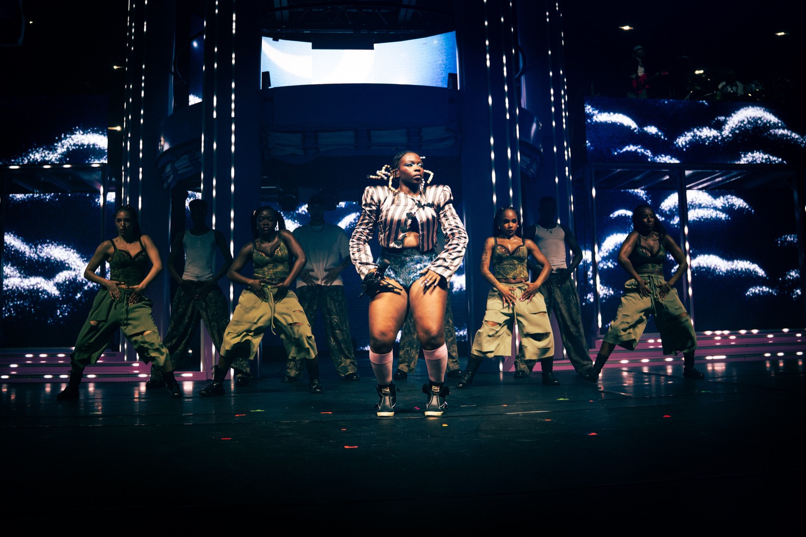 La chanteuse en short et blouson au centre de la scène, entourée de danseuses en combinaison kaki, sur fond de lumières bleues.