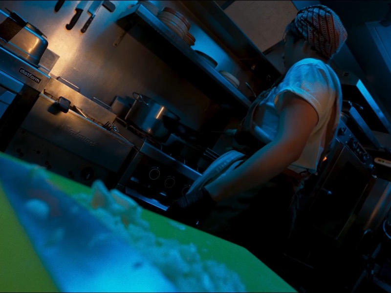 Dans une cuisine éclairée de bleu et de vert, une personne en bandana travaille devant les fourneaux, une planche de découpe au premier plan.