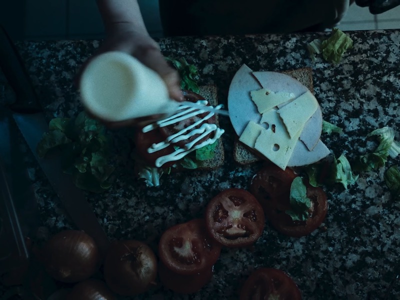 Vue plongeante sur deux mains qui versent une sauce sur un sandwich garni de salade et de fromage, des tomates coupées autour.