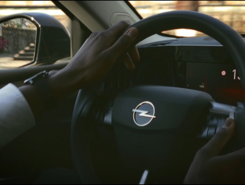 Gros plan sur deux mains posées sur un volant, le tableau de bord allumé et la rue défilant dans le rétroviseur.