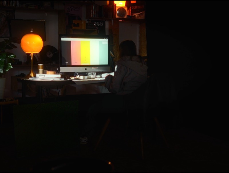 Dans une pièce sombre, une personne assise devant un grand écran affichant des barres de couleur, la lampe boule orange à gauche.